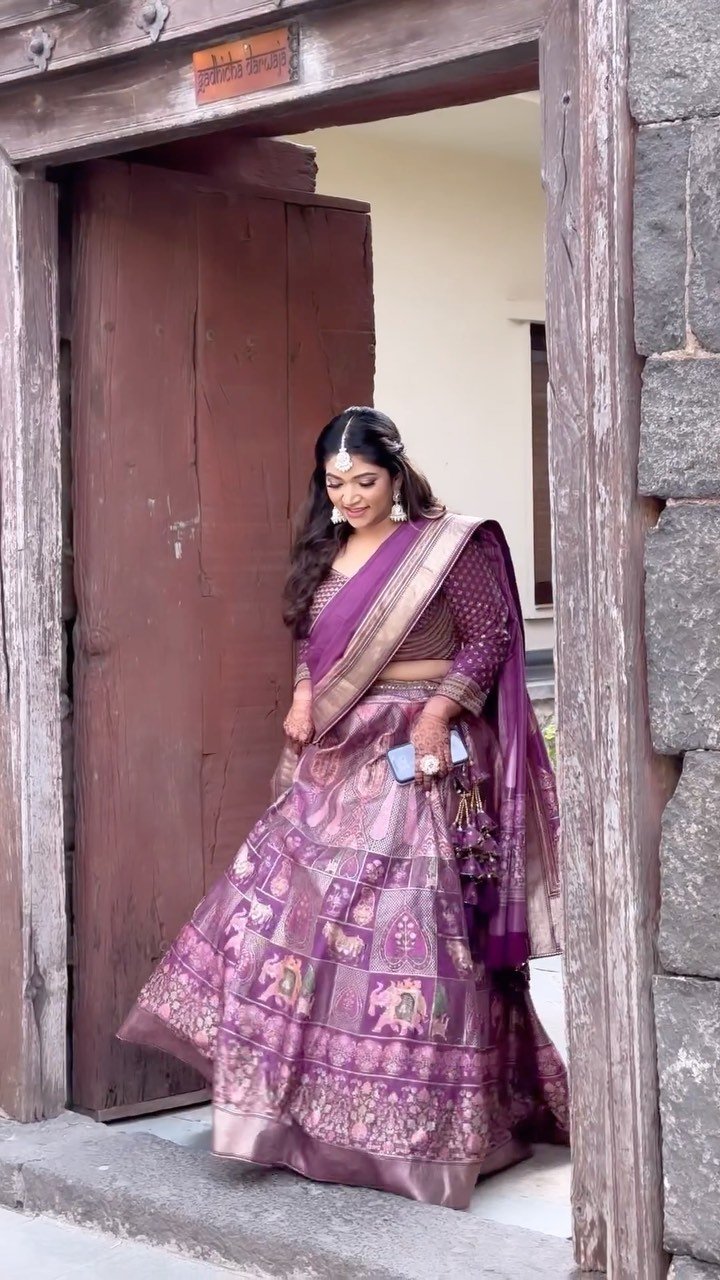 Bride in a lilac Banarasi lehenga photographed in a stone doorway at Fort JadhavGadh, Pune, makeup and hair by Tejaswini Gote
