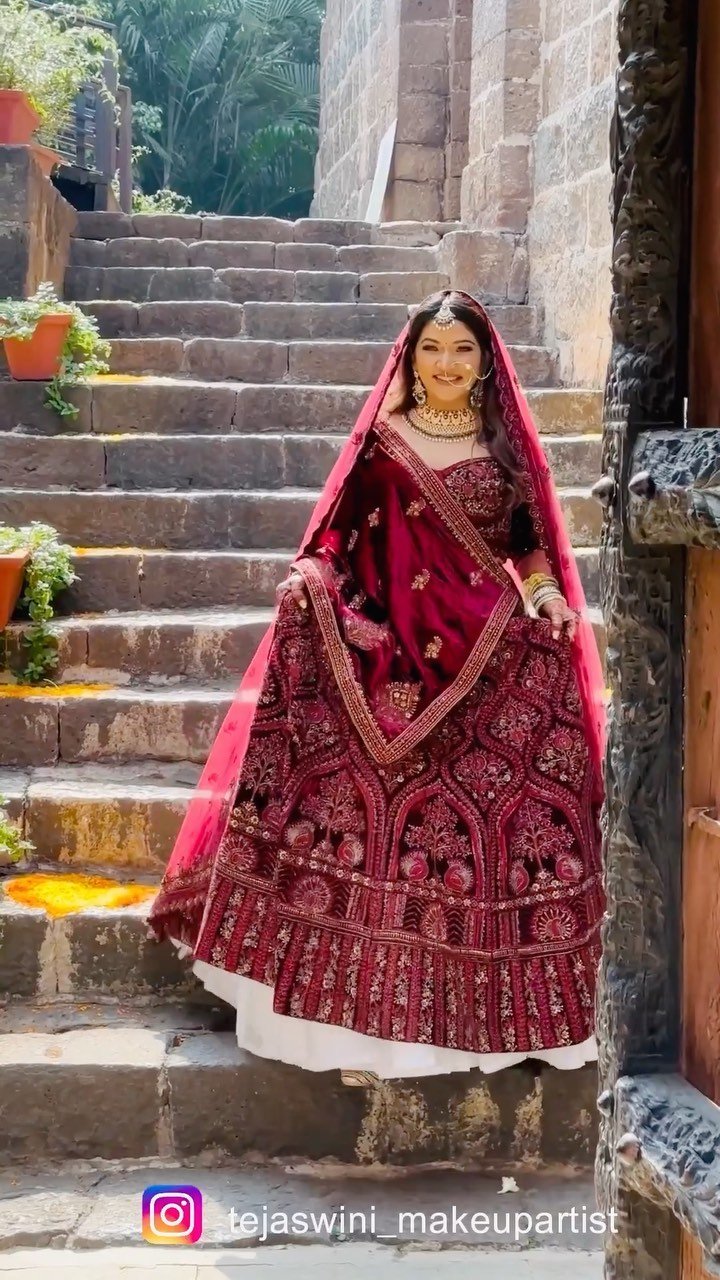Bride in a maroon lehenga on the stone steps at Fort JadhavGadh, Pune, bridal makeup, hair and draping by Tejaswini Gote